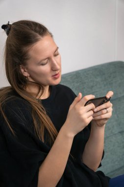 Düzgün giyimli genç bir kadın rahat bir koltukta oturuyor, evdeki mobil bir oyundan keyif alıyor, dijital eğlence ve eğlence dünyasına dalmış. Dikey fotoğraf