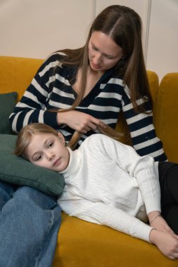 Anne, kızının yumuşak bir yastıkta dinlenirken nazikçe saçlarını okşuyor. Bu sırada her ikisi de, açık renkli sarı bir kanepede huzurlu bir günün tadını çıkarıyorlar. Dikey fotoğraf