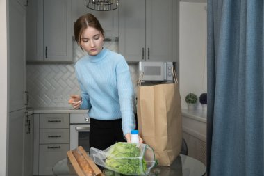 Mutfakta duran bir kız, marketten döndükten sonra masaya bir şişe süt koyuyor, masada bir kese kâğıt ve taze yiyecekle.