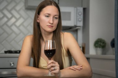 Mutfak masasında tek başına oturan genç bir kadın elinde bir kadeh kırmızı şarapla düşünceli ve yalnızlık, üzüntü ve duygusal karmaşa hislerini yansıtıyor.