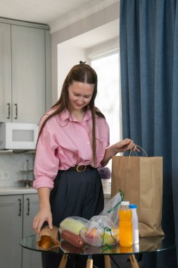 Genç bir kadın, modern bir mutfakta kese kağıdından yiyecek paketleri açıyor ve çevre dostu yeniden kullanılabilir poşetler açıyor, sürdürülebilir alışveriş uygulamalarını ön plana çıkarıyor ve sağlıklı yeme alışkanlıklarını teşvik ediyor. Dikey fotoğraf