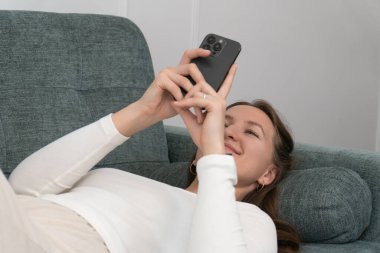 Evdeki rahat bir kanepede dinlenen genç bir kadın akıllı telefonuyla interneti ve sosyal medyayı karıştırıyor boş zamanlarının keyfini çıkartıyor.
