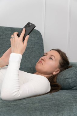 Genç kadın evinde rahat bir kanepede uzanıyor, akıllı telefon kullanıyor, internette geziniyor, sosyal medyada sohbet ediyor, video ve fotoğraf izliyor, üzgün bir yüz ifadesi takındı. Dikey fotoğraf