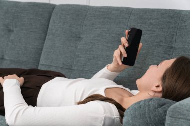 Genç kadın rahat bir şekilde kanepede uzanıyor, akıllı telefonunu kullanıyor, evde boş zamanlarının tadını çıkaran bir sesli mesaj kaydediyor, günlük rutinine ara veriyor.