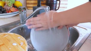 Kadının elleri kirli bulaşıkları dikkatlice yıkıyor. Tabak, bardak ve tahta kesme tahtası dahil. Evdeki metal mutfak lavabosunda musluk suyunun altında bir sünger ve sabun var.