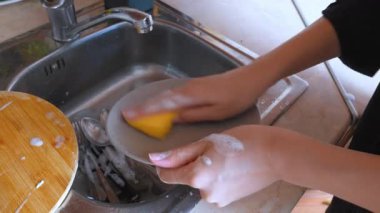 Kadın elleri kirli bir bulaşığı paslanmaz çelik bir lavaboda sabunlu bir süngerle güzelce yıkıyor. Günlük ev işlerini gösteriyor ve mutfağı temiz tutuyor.