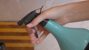 Bir kadının elinin turkuaz plastik bir şişeyi tutarken yakın plan görüntüsü ve su püskürtmek için defalarca tetiğe basması temizlik, bahçe işleri veya ev işlerini sembolize ediyor.