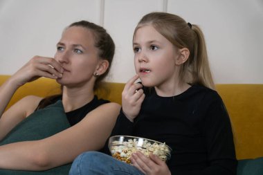 Anne ve kızı evde sarı bir kanepede dinleniyor, patlamış mısırlarını paylaşıyorlar ve samimi bir aile filmi gecesi boyunca birlikte film izliyorlar.