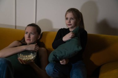 İki kadın, koyu renkli bir odada, sarı bir kanepede oturur. Bir yetişkin, elinde patlamış mısır, elinde yastık tutarken korkmuş bir şekilde bakar. İkisi de yoğun bir film izler.