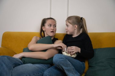 İki genç kız kardeş sarı bir kanepede oturmuş korkunç bir film izlerken patlamış mısırlarını paylaşıyorlar. Bir kız kardeş korkmuş görünüyor, diğeri de ona bakıyor.