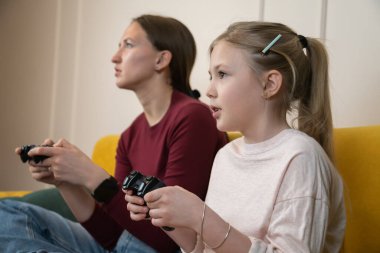Anne ve kız evde oturuyor, kontrolörlerle oyun oynarken gülümsüyor ve konsantre oluyor, eğlenceli bağları, boş vakitleri ve modern aile eğlencelerini paylaşıyorlar.