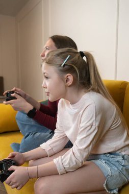 Konsantre anne-kız video oyunu oynarken birbirlerine bağlanıyorlar, kumandayı tutuyorlar ve sarı bir kanepede oturup evde boş vakitlerinin tadını çıkarıyorlar. Dikey fotoğraf