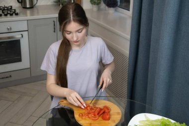 Genç kız, ahşap bir kesim tahtasıyla taze domatesleri dikkatlice dilimliyor, modern mutfak ortamında sağlıklı bir yemek veya salata hazırlıyor, yanında da marul var.