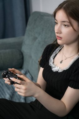 Oyun kumandası tutan genç bir kız oturma odasında rahat bir kanepede oturmuş video oyunları oynuyor ve boş zamanlarının tadını çıkarıyor. Dikey fotoğraf