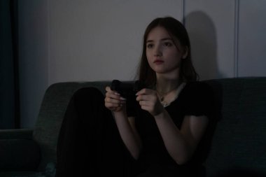 Genç bir kız, elinde bir video oyun kumandası, koltukta rahat bir şekilde otururken bir anlık rahatlığın tadını çıkarıyor. Sahne, konforlu bir ortamda eğlence ve eğlence sunar.