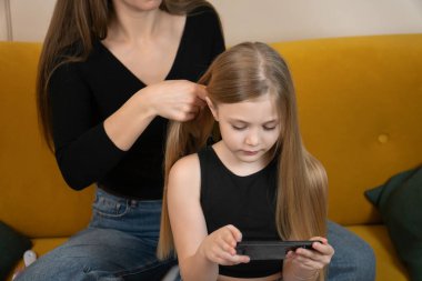 Anne kızının uzun sarı saçlarını örerken çocuk akıllı telefona odaklanıyor, sıcak bir aile bağı kurma anı yakalıyor, çoklu görev ve günlük ev rutini.