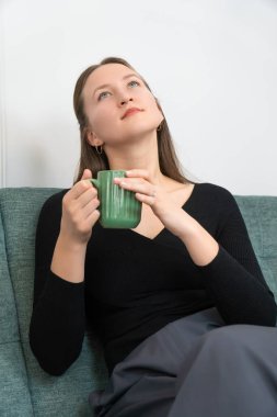 Kanepede rahat bir şekilde oturan genç bir kadın, elinde yeşil bir kupayla, düşünceli bir tefekküre dalmış, huzur ve huzur dolu bir anın tadını çıkarıyor. Dikey fotoğraf