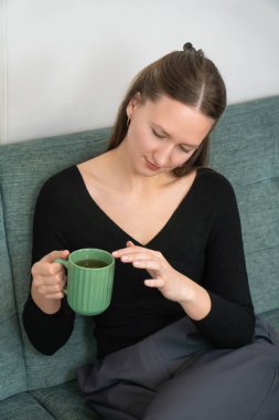 Genç bir kadın rahat bir şekilde kanepede oturur, sıcak bir fincan bitki çayıyla huzurlu bir anın tadını çıkarır, rahat ve huzurlu bir atmosfer yaratır. Dikey fotoğraf