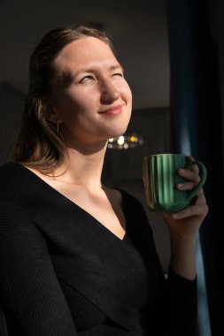 Kapalı gözleri ve sakin bir ifadeyle, genç bir kadın sabah içeceğinin sıcaklığının tadını çıkararak, yakınlardaki bir pencereden süzülen yumuşak güneş ışığının tadını çıkarıyor. Dikey fotoğraf