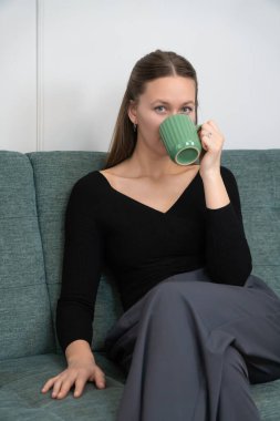 Genç bir kadın rahat bir kanepede uzanırken yeşil bardaktan sıcak bir içecek yudumluyor, rahat ve rahatlatıcı bir atmosfer yaratıyor rahatlama ve keyif için mükemmel. Dikey fotoğraf