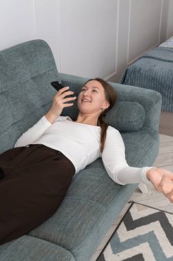 Genç kadın rahat bir şekilde kanepede uzanıyor, akıllı telefonunu kullanıyor, evde boş zamanlarının tadını çıkaran bir sesli mesaj kaydediyor, günlük rutinine ara veriyor. Dikey fotoğraf