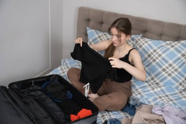 Genç kız bir yatakta oturuyor, giysileri katlıyor ve açık bir bavul hazırlıyor, yaklaşan bir yolculuk için valizleri ve gerekli şeyleri hazırlıyor ya da sıcak bir yatak odasında tatil yapıyor.