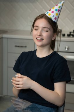 Genç bir kız mutfak masasında oturuyor, gözleri kapalı ve renkli bir doğum günü şapkası takıyor, bir dilek dilerken evinde mutlu bir şekilde kutluyor.