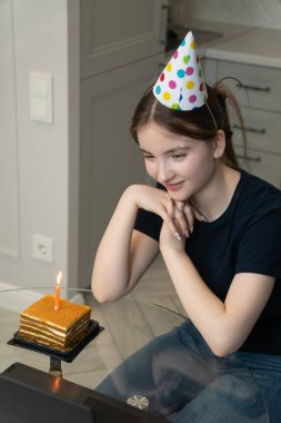 Young woman in party hat making video call on laptop, celebrating her birthday online with friends and family during quarantine or living apart