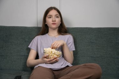 Comfortable girl enjoying a movie night, eating popcorn from a bowl while relaxing on a sofa in her living room, with the remote control nearby