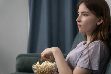 Genç kız rahat bir koltukta oturup televizyon izliyor ve sessiz oturma odasında cam kaseden patlamış mısır yiyor, evinde sakin bir akşamın tadını çıkarıyor.