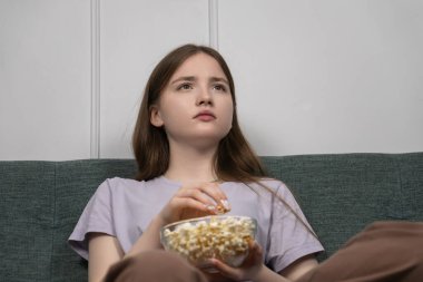 Genç kız rahat bir kanepede oturmuş, bir kase patlamış mısırla film gecesinin tadını çıkarıyor, evde boş vakit ve eğlence anı yaşıyor.