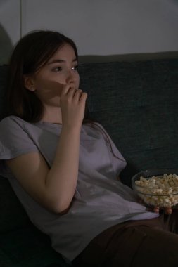 Rahat bir genç kız evinde rahat bir akşamın tadını çıkarıyor, kanepede oturuyor, televizyon izliyor ve yanında uzaktan kumandayla cam bir kaseden patlamış mısır yiyor.