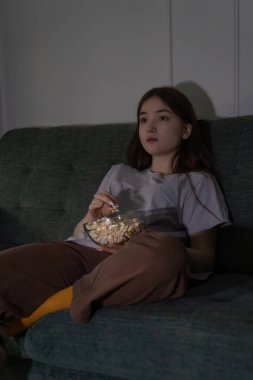 Düşünceli genç kız televizyon seyredip patlamış mısırın tadını çıkarırken loş ışıklı bir oturma odasında dinleniyor ve evdeki sessiz bir gecede huzurlu bir atmosfer yaratıyor.