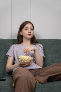 Rahat genç kız kanepede rahat bir film gecesinin tadını çıkarıyor, büyüleyici bir televizyon şovuna dalmışken cam kaseden patlamış mısır yiyor. Dikey fotoğraf