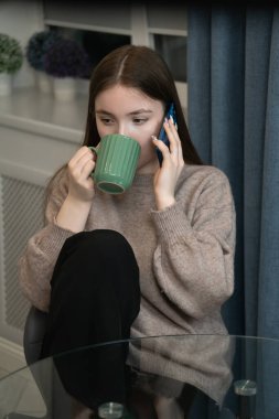Young girl enjoys a relaxing moment at home, sitting comfortably while sipping tea from a green mug and engaging in a phone conversation