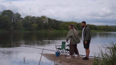 Genç çift, sakin bir günün tadını çıkarırken, bir adam sabırla kadınlara sakin bir nehrin kumlu sahiline olta atmayı öğretiyor.
