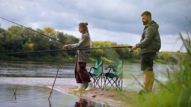 Millennial erkek ve kadın rahat bir günün tadını çıkarıyorlar, kumlu bir nehir kıyısında duruyorlar ve balıkçılık malzemelerini doğada eğlenceli bir balık tutma seansı için birlikte hazırlıyorlar.