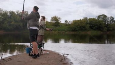 Genç bir adam ve kadın kumlu bir nehir kıyısında durup olta kamışı tutarken ve sabırla avlanmak için beklerken açık hava aktivitelerinin ve birlikte sakin bir anın tadını çıkarıyorlar.
