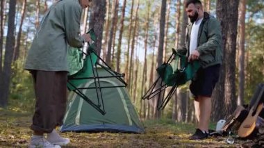 Beyaz erkek ve kadın, güneşli bir çam ormanında çadırlarının yanına sandalye ve masa kurarak kamplarını yakınlarda bir gitarla dinlendirici bir geziye hazırlıyorlar.