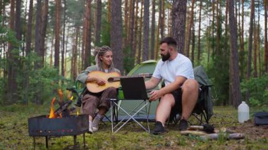 Genç hippi kadın, erkek arkadaşı çadır ve şenlik ateşinin yanında dizüstü bilgisayarda çalışırken gitar çalıyor. Romantik bir kaçamağın ve güzel bir çam ormanında uzaktan çalışmanın keyfini çıkarıyor.