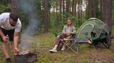 Genç bir adam barbeküde et pişirirken kız arkadaşı da bir çadırı olan kamp alanında oturuyor. Çift, güzel bir çam ormanında huzurlu bir pikniğin ve açık havada eğlencenin tadını çıkarıyor.