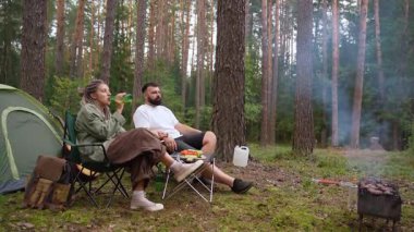 Genç heteroseksüel çift çadırın yanında kamp sandalyelerinde oturuyor ve güzel bir çam ormanında kamp ateşi yakıyor, bira içen adam ve sakin akşam atmosferinin tadını çıkaran kadın.