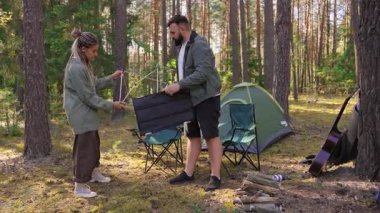 Genç bir çift, kamp malzemeleri, çadır ve uzun ağaçlarla çevrili güneşli bir çam ormanında kamp alanlarında portatif bir katlama masası kurmak için birlikte çalışıyor.