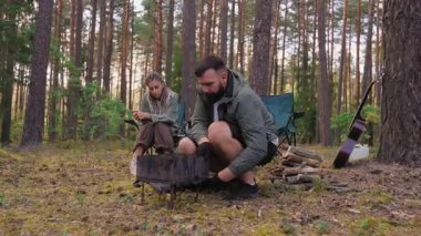 Genç bir adam piknik için barbekü ızgarası hazırlarken bir kadın katlanan sandalyede oturuyor. Çift güzel bir çam ormanında çadır ve gitarla romantik bir hafta sonu geçiriyor.