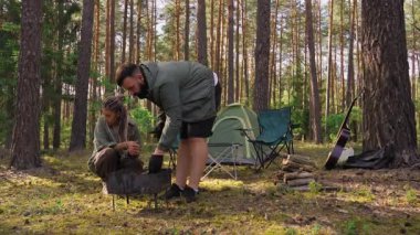 Genç bir adam piknik için barbekü ızgarası hazırlarken bir kadın katlanan sandalyede oturuyor. Çift güzel bir çam ormanında çadır ve gitarla romantik bir hafta sonu geçiriyor.