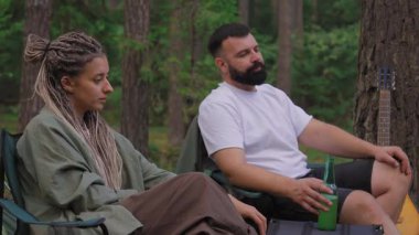 Rahat genç bir çift kamp sandalyelerinde oturuyor, konuşuyor ve güzel bir çam ormanında piknikte eğleniyorlar. Arka planda bir ağaca yaslanmış bir gitarla.