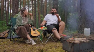 Rastalı genç bir kadın salata için taze sebze dilimlerken erkek arkadaşı da güzel yeşil bir çam ormanında kamp yaparken kamp ateşinde barbekü izliyor.