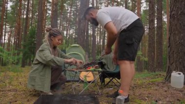Mutlu genç çift, yaz tatilleri boyunca çam ormanının ortasında pikniğin tadını çıkarırken ızgarada ızgara ızgarasında leziz et pişiriyor.