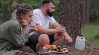 Rastalı ve sakallı beyaz bir çift ormanda ızgara et ve taze sebzelerle pikniğin tadını çıkarıyor. Seyahat, turizm ve açık hava maceralarını tanıtıyorlar.