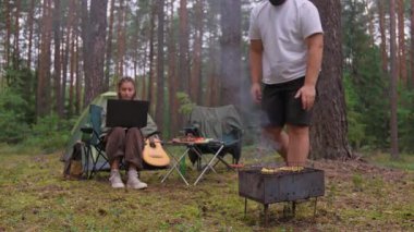 Genç bir kadın laptopunun bilgisayarında çalışırken partneri de güzel bir çam ormanındaki kamp gezisi sırasında barbeküde yemek pişiriyor. İş ve boş zamanı doğada birleştiriyor.